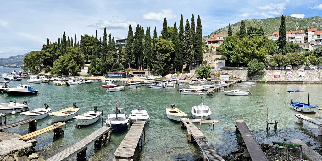 Small boats moored at wooden jetties in Cavtat harbour, with cypress trees and houses behind
