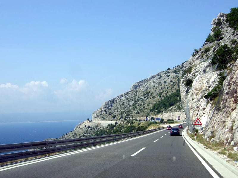 Cars on a coastal road cut into limestone cliffs above the Adriatic, with a tunnel ahead